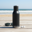 Asobu wireless Bluetooth speaker bottle on a sunny beach boardwalk with ocean in the background